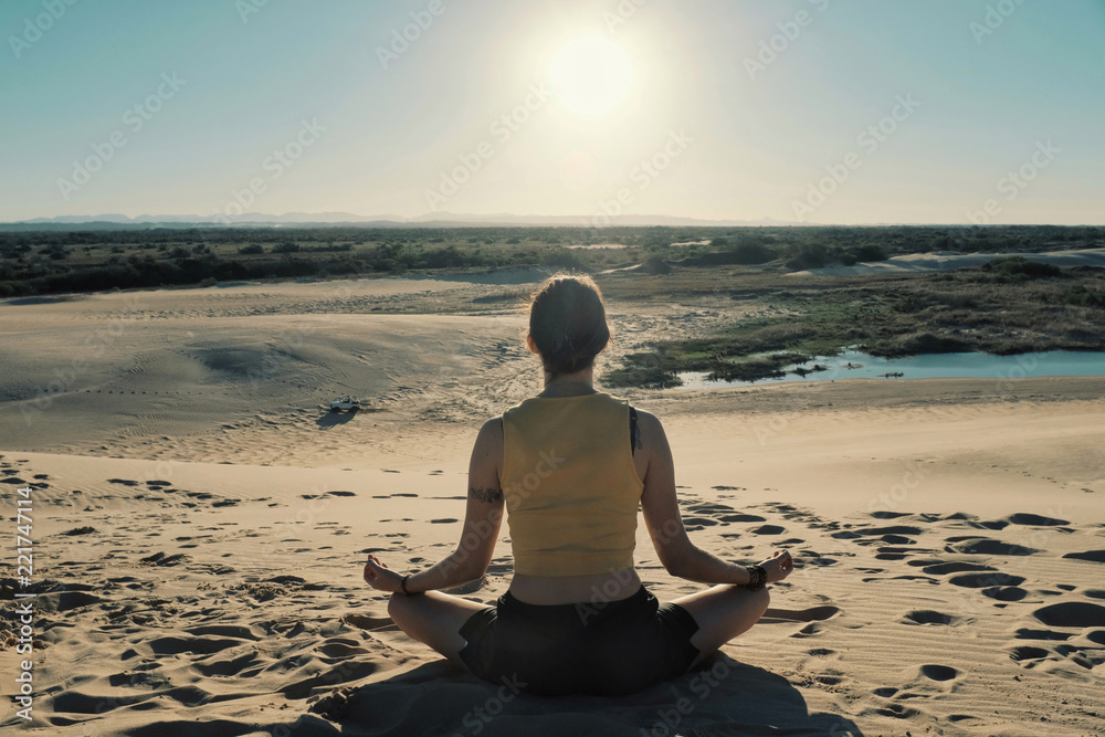 young woman sitting in a meditation mantra yoga pose on the top of a desert sand dune in front of a lagoon lake at sunset