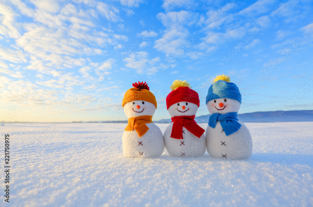 Naklejka premium Friends snowman in red, blue, yellow hats and scarfs. Nice winter scenery with mountains, field in snow. Orange sunset background.