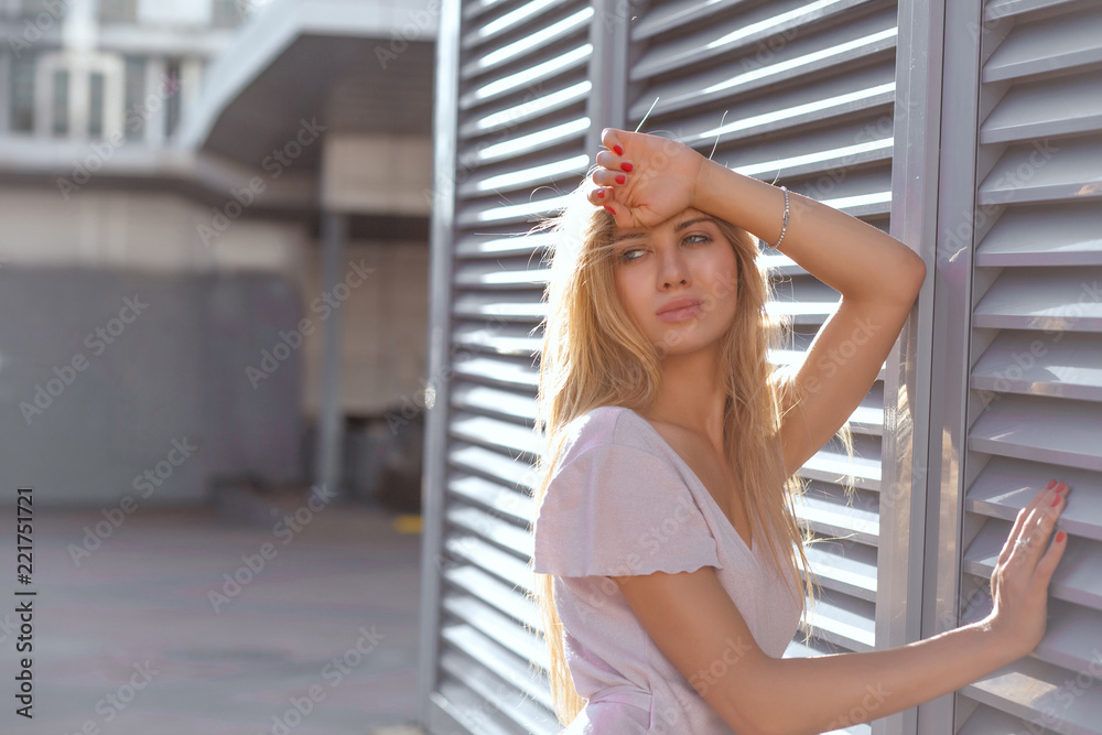Stunning blonde girl with long hair posing at the shutters with sun glare. Space for text