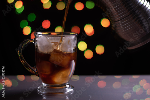 Glass With Ice And Coffee Poured Into It By A Stainless Steel Coffee Maker On A Shiney Platform With Dark Background And Colorful Bokeh Lights photo