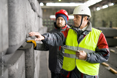 Waist up portrait of mature worker  wearing warm jacket and hardhat using digital tablet while discussing concrete blocks production with trainee in workshop photo