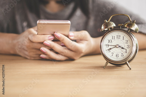 wasting time at work concept with woman using smart phone with alarm clock on desk