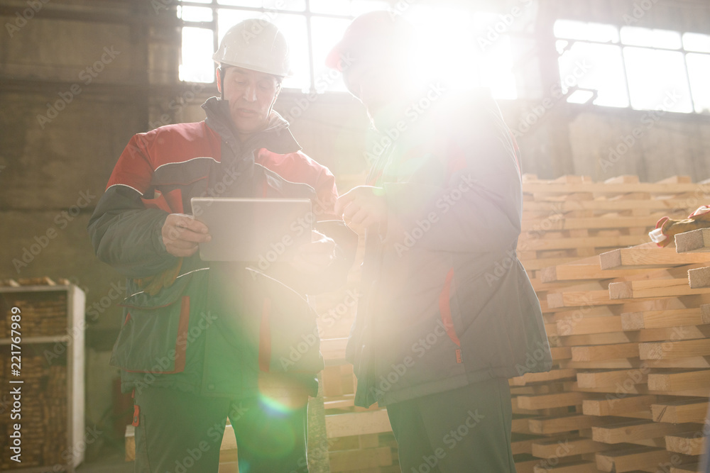 Waist up portrait of two factory workers using digital tablet and  discussing production process while standing in sunlight