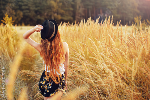 Beauty Girl Outdoors enjoying nature. Pretty Teenage Model in hat running on the Spring Field, Sun Light. Romantic young blonde girl in a wheat field. Woman in Casual. Blowing Long Hair. Sunset.