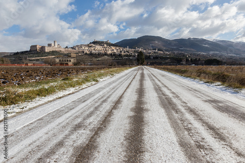 View of Assisi town (Umbria) in winter, with a country road covered by snow and a blue sky with white clouds