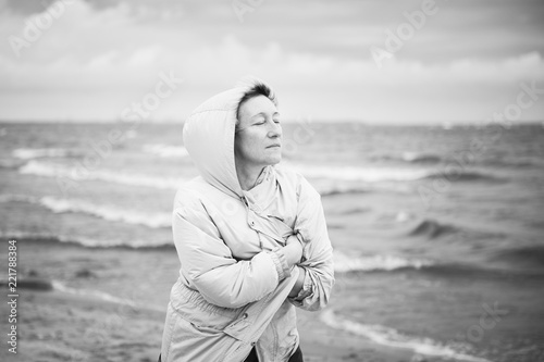 Beautiful adult female wrapping in warm jacket and keeping eyes closed while walking near waving sea