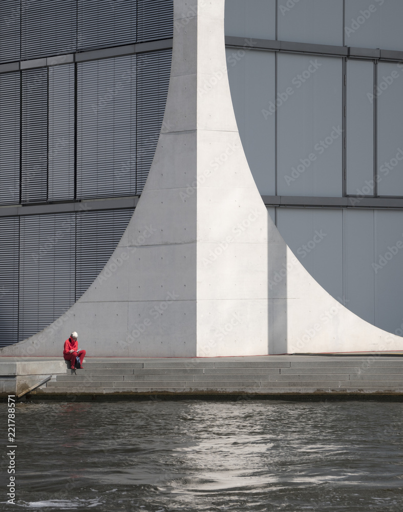 anonymous woman in red sits on the river Spree in front of the Marie-Elisabeth-Lueders-Haus, a modern building made of concrete and glass in Berlin anonymous woman in red sits on the river Spree in front of the Marie-Elisabeth-Lueders-Haus, a modern building made of concrete and glass in Berlin
