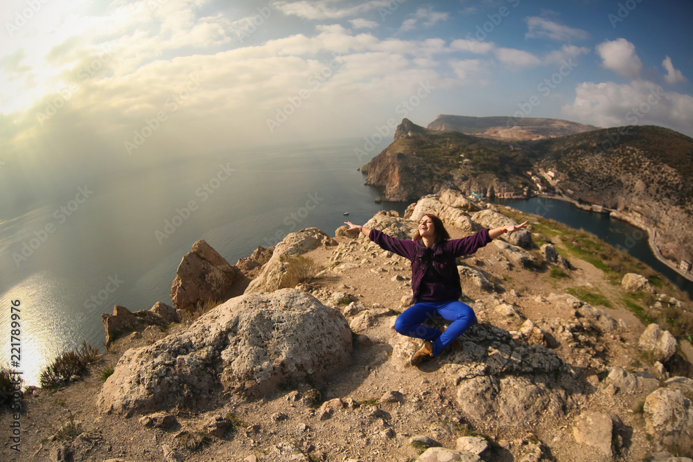 Beautiful young happy carefree woman traveler sits on top of a hill with a stunning amazing sea view, with arms outstretched