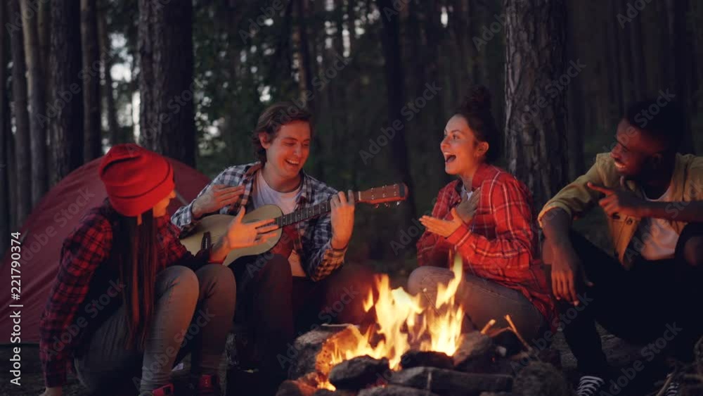 Cinemagraph loop - young man tourist is playing the guitar while his friends are singing and laughing sitting around fire in the wood in the evening enjoying nature and company.