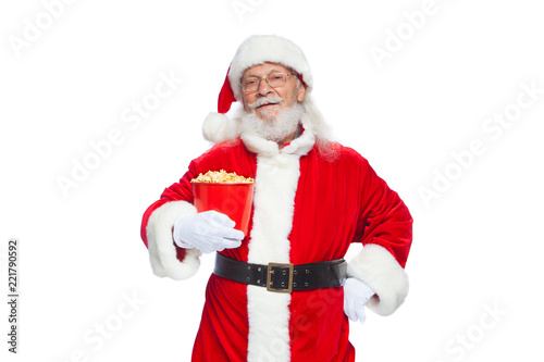 Christmas. Smiling, kind Santa Claus in white gloves holds a red bucket with popcorn with one hand, and puts the other hand on his belt. The concept of visiting the cinema, watching a movie with