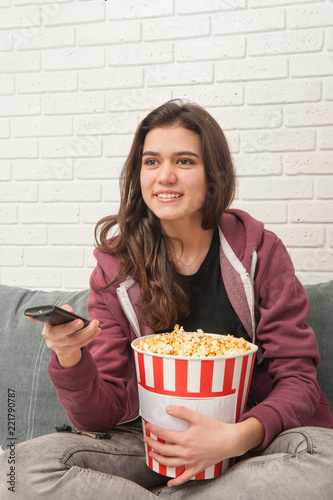 Teen girl sitting on couch with TV remote and eating popcorn
