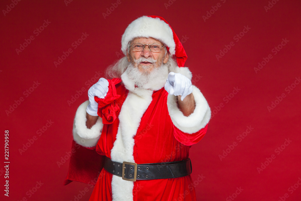 Christmas. Santa Claus in white gloves with a dissatisfied face expression with a bag of gifts behind his back points his index finger into the camera. Isolated on red background.