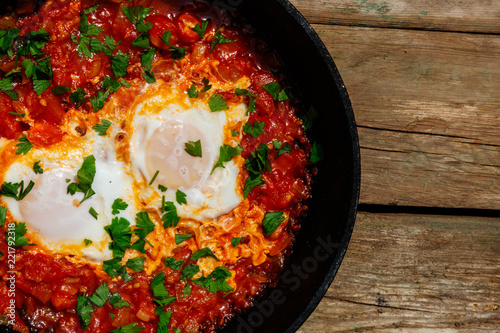 Homemade breakfast shakshuka - fried eggs, onion, bell pepper, tomatoes and parsley in cast-iron frying pan on rustic wooden table. Jewish cuisine. Top view