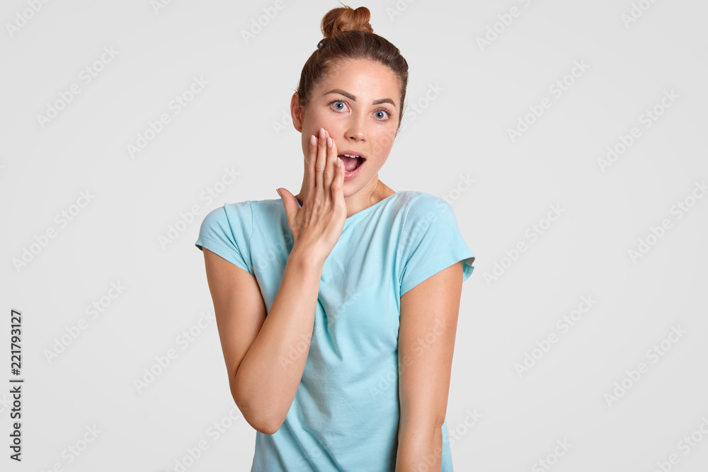 Photo of amazed fit girl keeps jaw dropped, touches cheek with hand, has bun knot, dressed in comfortable light blue t shirt, wonders latest news, isolatedd over white studio wall. Emotions concept Photo of amazed fit girl keeps jaw dropped, touches cheek with hand, has bun knot, dressed in comfortable light blue t shirt, wonders latest news, isolatedd over white studio wall. Emotions concept