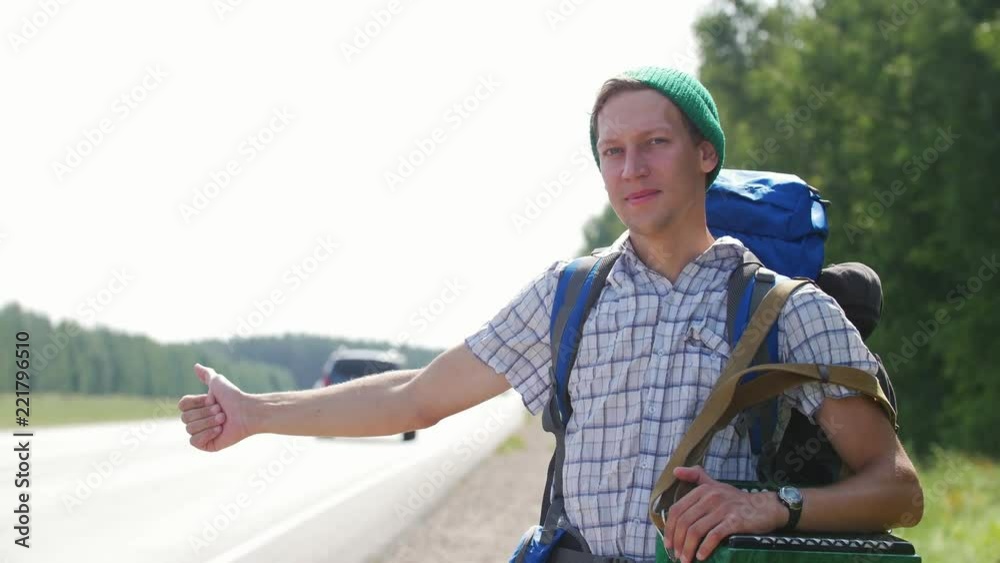 A young man in a plaid shirt, a green hat and a backpack standing by the road and honking cars A young man in a plaid shirt, a green hat and a backpack standing by the road and honking cars