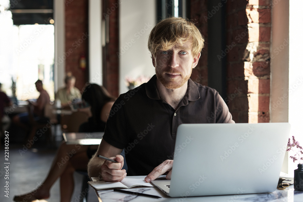 Handsome ginger caucasian man writing in notebook using laptop while sitting in cafe having coffee. Concept of young business people working in public space or co-working. Handsome ginger caucasian man writing in notebook using laptop while sitting in cafe having coffee. Concept of young business people working in public space or co-working.
