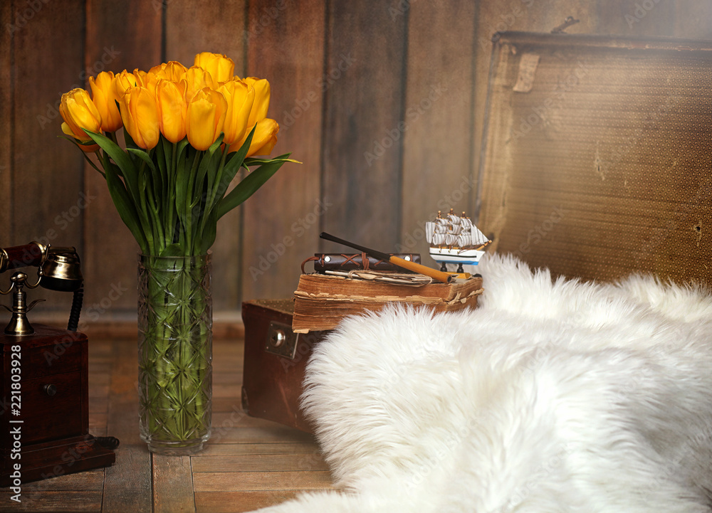 A bouquet of yellow tulips in a vase in the interior of a retro room. Retro interior with bouquet of tulips old phone and suitcase. A bouquet of yellow tulips in a vase in the interior of a retro room. Retro interior with bouquet of tulips old phone and suitcase.