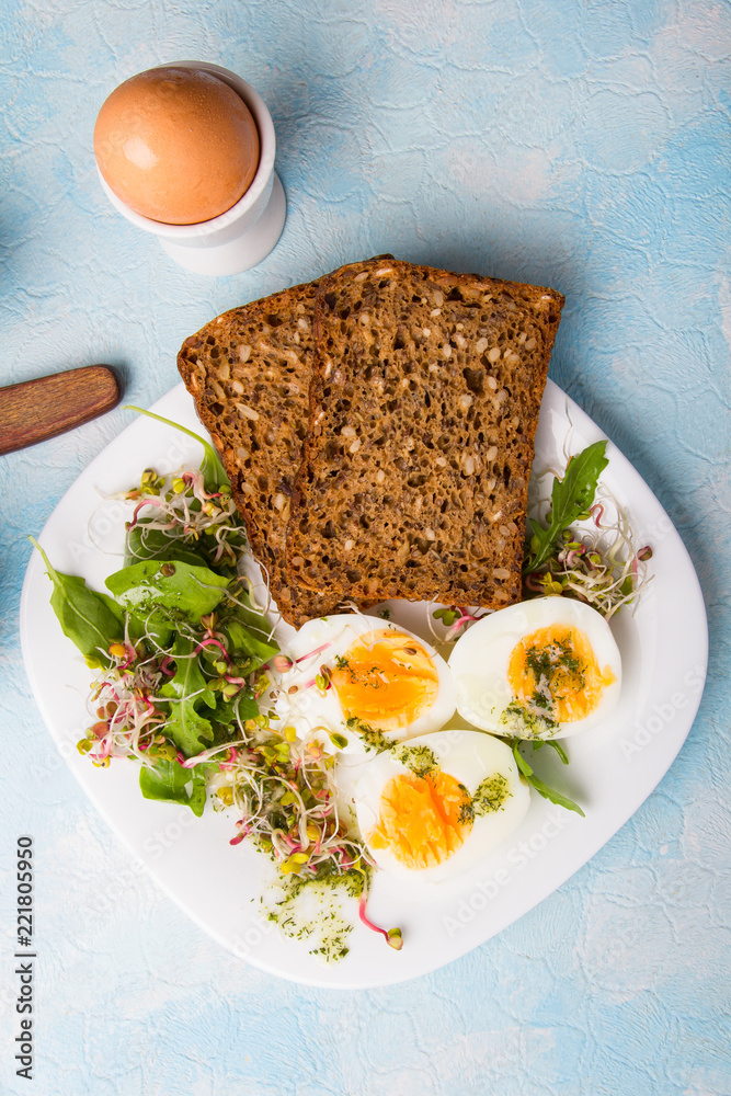Healthy breakfast: Hard boiled eggs, fresh radish sprouts, arugula and dark whole wheat  bread with herb sauce, on blue background