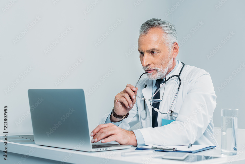 middle aged male doctor holding eyeglasses and using laptop at table in office middle aged male doctor holding eyeglasses and using laptop at table in office