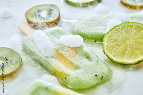 Melting green ice cream on a stick with pieces of peach on a gray background with pieces of ice, kiwi, lime and kiwi. Summer dessert