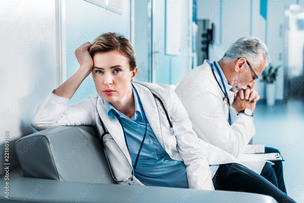tired female doctor looking at camera on sofa while her male colleague sitting behind in hospital corridor
