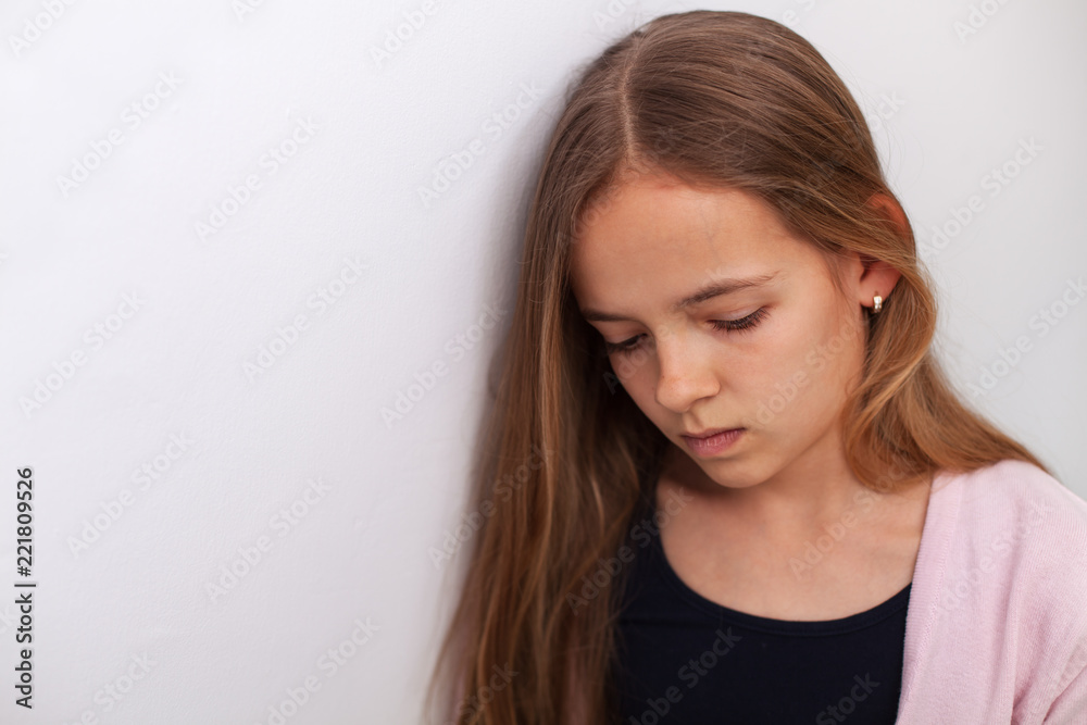 Sad teenager girl standing by the white wall with downcast eyes Sad teenager girl standing by the white wall with downcast eyes