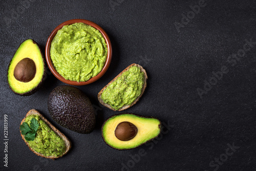 Traditional Mexican Dip Sauce Guacamole in a bowl with bread toasts,  whole and cut half avocado  on dark background. Top view. Copy space.