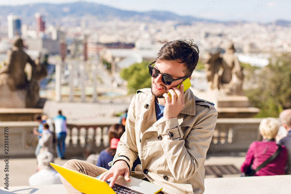 Portrait of young guy in beige coat sitting on hight on city background. He wears beige coat, working on yellow laptop, speaking on phone.