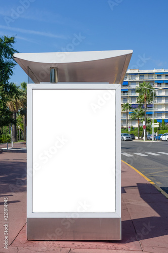 Vertical blank white billboard at bus stop on city street. In the background buildings and road. Mock up. Poster on street next to roadway. Sunny summer day. Cannes, France © florent