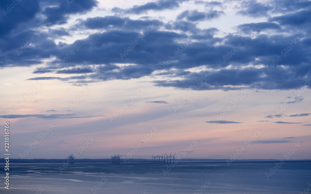 Sea view of the beach at sunset in the autumn with orange,purple sky with dramatic dark blue clouds,Relaxing waves rolling in with the gentle tide,Roll of windmills standing in the sea on the horizon