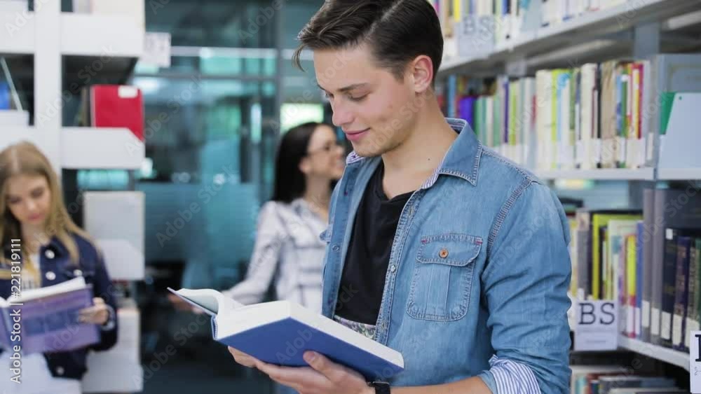 Library. Smiling Male Student Reading Book Near Shelves Library. Smiling Male Student Reading Book Near Shelves