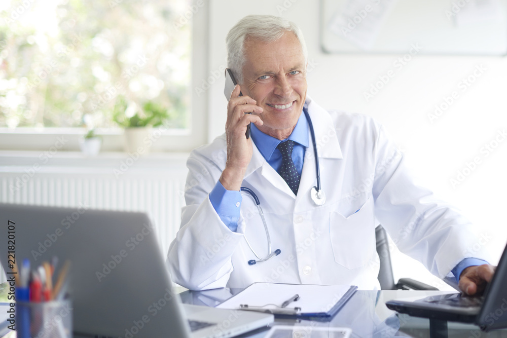 Male doctor making call. Shot of a doctor sitting at consulting room and talking with his patient on phone. 