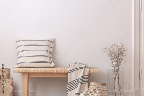 Pillow and blanket on wooden bench next to flowers in beige minimal flat interior. Real photo