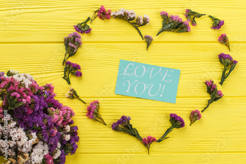 Fototapeta Naklejka Na Ścianę i Meble -  Love you concept with flowers, flat lay. Statice limonium flowers forming heart shape and paper with love you text. Yellow wooden table surface background.