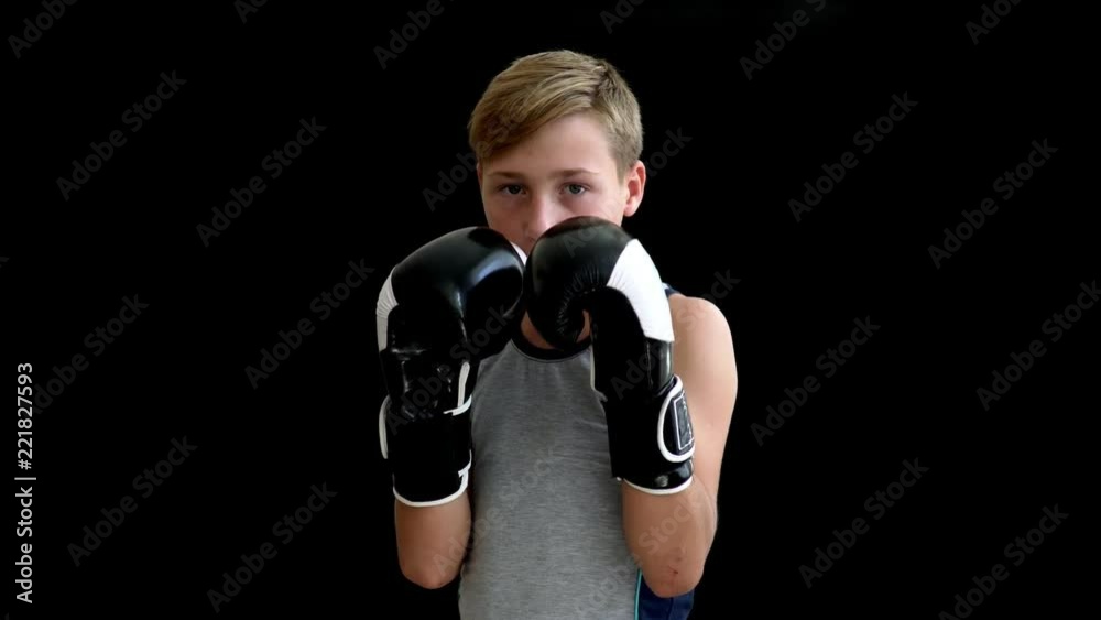 A teenage boxer with blond hair and dark eyes stands on a dark background. Hands of a teenager descend along the body, on it - a gray shirt without sleeves, a dark background.