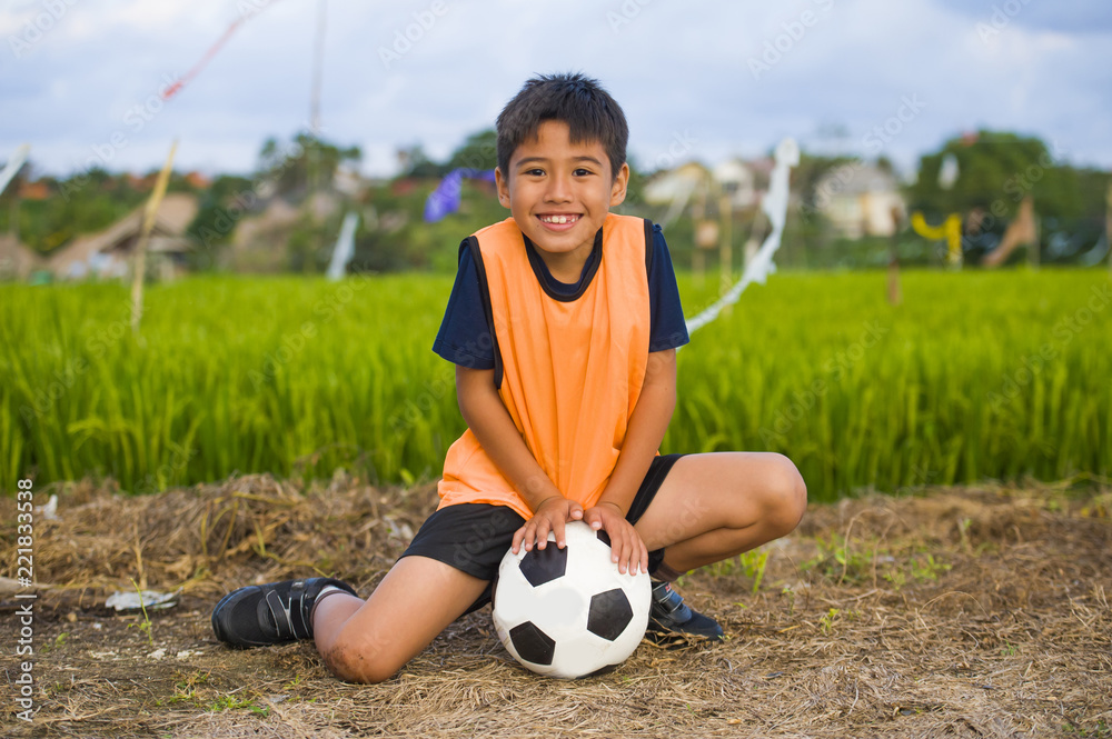 lifestyle portrait of handsome and happy young boy holding soccer ball playing football outdoors at green grass field smiling cheerful wearing training vest