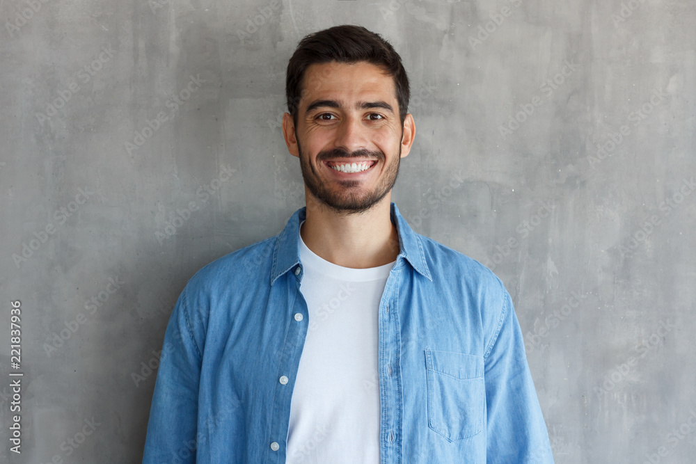 Horizontal closeup of young handsome European Caucasian male isolated on grey background wearing blue denim shirt and white T-shirt, having short and face hair, smiling confidently and friendly Horizontal closeup of young handsome European Caucasian male isolated on grey background wearing blue denim shirt and white T-shirt, having short and face hair, smiling confidently and friendly