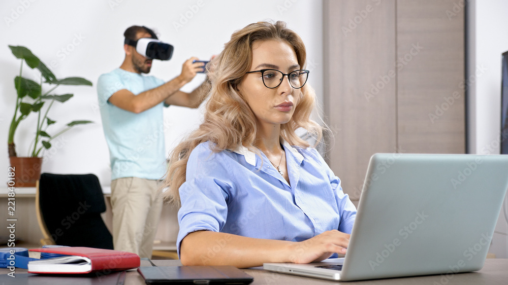 Tired and upset freelancer woman working on the computer laptop in the house while her husband is disturbing her by playing video games with a VR headset in the background. Tired and upset freelancer woman working on the computer laptop in the house while her husband is disturbing her by playing video games with a VR headset in the background.