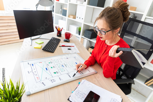 A young girl sits at a table in the office, holds a marker in her hand and works with a notepad and a magnetic board. photo