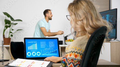 Freelancer woman analysing business charts on the computer while her husband is playing video games on the TV in background