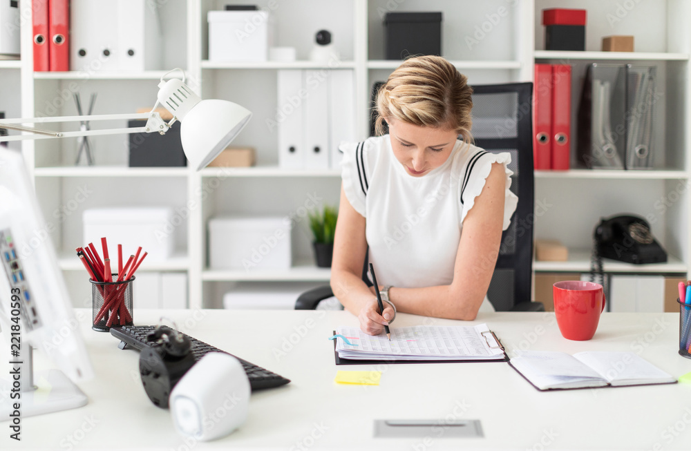 A young blonde girl sits at a computer desk in the office and writes a pencil in the document. A young blonde girl sits at a computer desk in the office and writes a pencil in the document.
