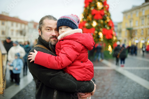 Adorable girl and her father having wonderful time on traditional Christmas market. Parent and child enjoying themselves near Christmas stands decorated with lights and baubles.