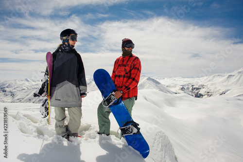 guy and a girl in warm clothes stand with snowboards against the mountain snow tops