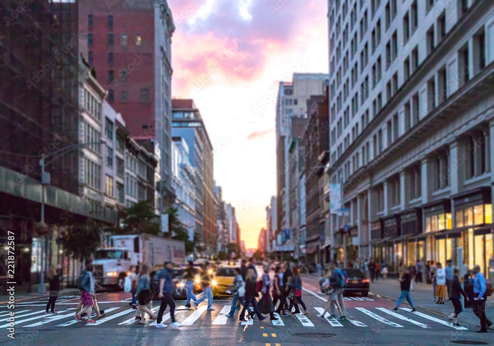 Diverse group of people walking across the busy intersection 23rd Street and 6th Avenue in Manhattan with the colorful light of sunset shining through the skyline buildings