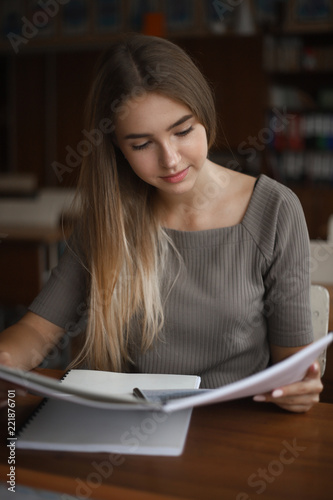 Concentrated beautiful young student schoolgirl lady with long hair sitting at the table in classroom with copybook reading.