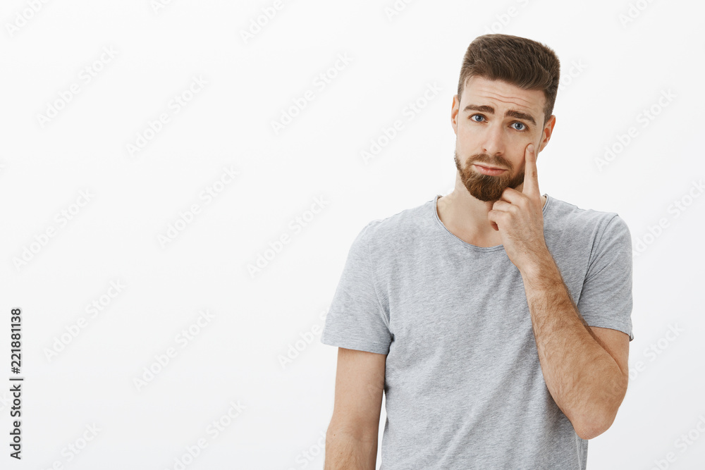 Handsome young touched guy with beard making sad and gloomy expression pointing at eyelid as if showing teardrop expressing regret or sadness standing displeased crying over gray background Handsome young touched guy with beard making sad and gloomy expression pointing at eyelid as if showing teardrop expressing regret or sadness standing displeased crying over gray background