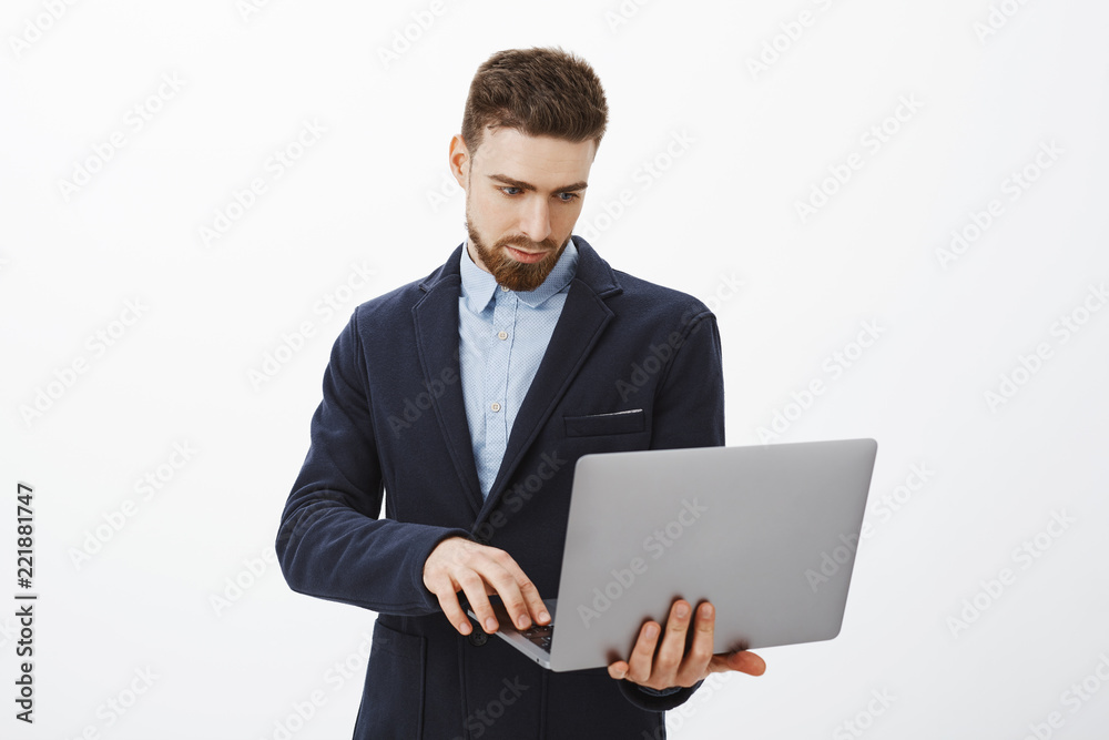 Focusing on business. Portrait of smart and ambitious concentrated good-looking young male entrepreneur with beard and blue eyes holding laptop in hand browsing checking schedule with determined look Focusing on business. Portrait of smart and ambitious concentrated good-looking young male entrepreneur with beard and blue eyes holding laptop in hand browsing checking schedule with determined look