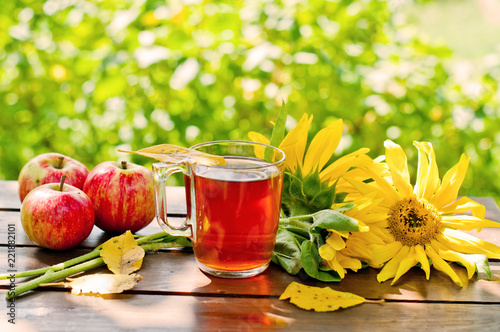Autumn background, hot tea in a glass and yellow sunflowers with apples on a wooden table in the garden.