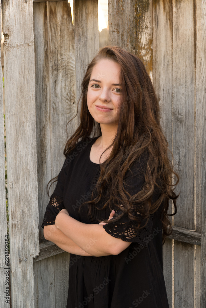 Brunette woman posing in front of a rustic old wooden fence wearing a black dress standing with arms crossed. Portrait of a young white woman standing in front of a wood fence outside in the summer.