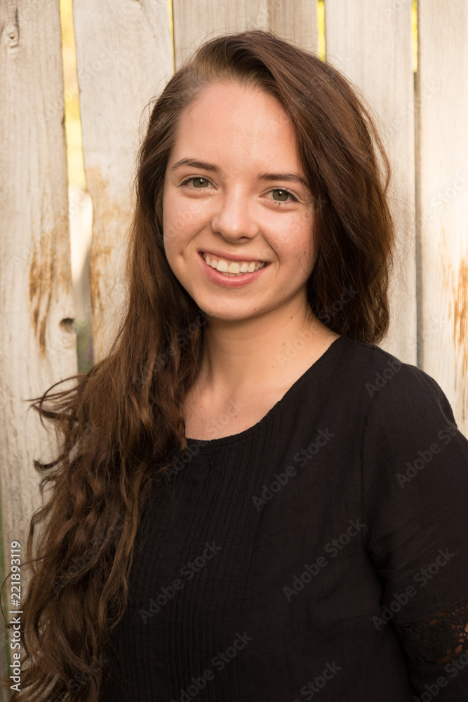 Portrait of a young Caucasian woman with long brown hair wearing a black dress standing next to a rustic old wooden fence. Model head shot of young woman, brunette hair. Soft natural light. Portrait of a young Caucasian woman with long brown hair wearing a black dress standing next to a rustic old wooden fence. Model head shot of young woman, brunette hair. Soft natural light.